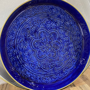Puede incluir: Una bandeja redonda de cerámica azul oscuro con un detallado diseño floral. La bandeja tiene un borde dorado y un diseño en relieve texturizado. La bandeja está sobre una superficie de madera.