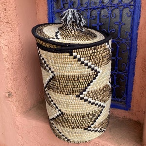 May include: A woven storage basket with a lid. The basket is cylindrical with a black rim and a decorative pattern in black, white, and tan. The lid has a tassel on top. The basket is sitting on a pink surface.
