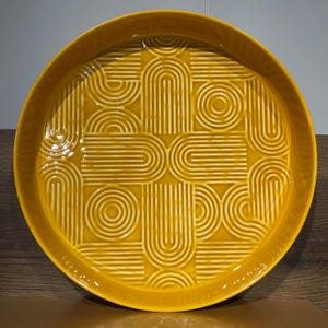 May include: A round, mustard-yellow ceramic serving tray with a raised rim. The tray features a repeating geometric pattern of curved lines and circles, creating a textured surface. The design is a lighter shade of yellow than the tray's base color.