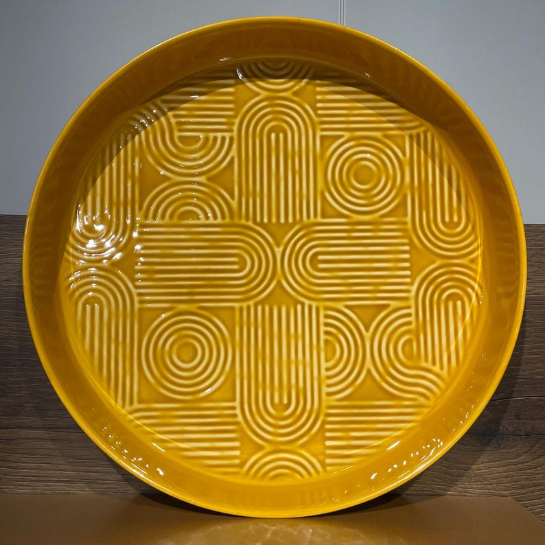 May include: A round, mustard-yellow ceramic serving tray with a raised rim. The tray features a repeating geometric pattern of curved lines and circles, creating a textured surface. The design is a lighter shade of yellow than the tray's base color.