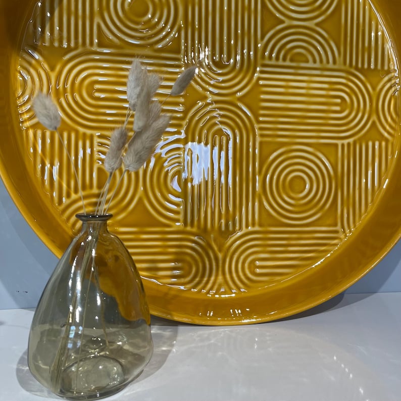 May include: A decorative still life featuring a yellow ceramic tray with a geometric pattern and a small, clear glass vase holding dried flowers. The tray is round and has a raised edge.