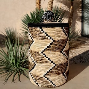 May include: Woven storage basket with a lid. The basket features a geometric pattern in shades of beige, brown, and black. The lid is black with a decorative tassel. The basket is cylindrical and appears to be made of natural fibers.