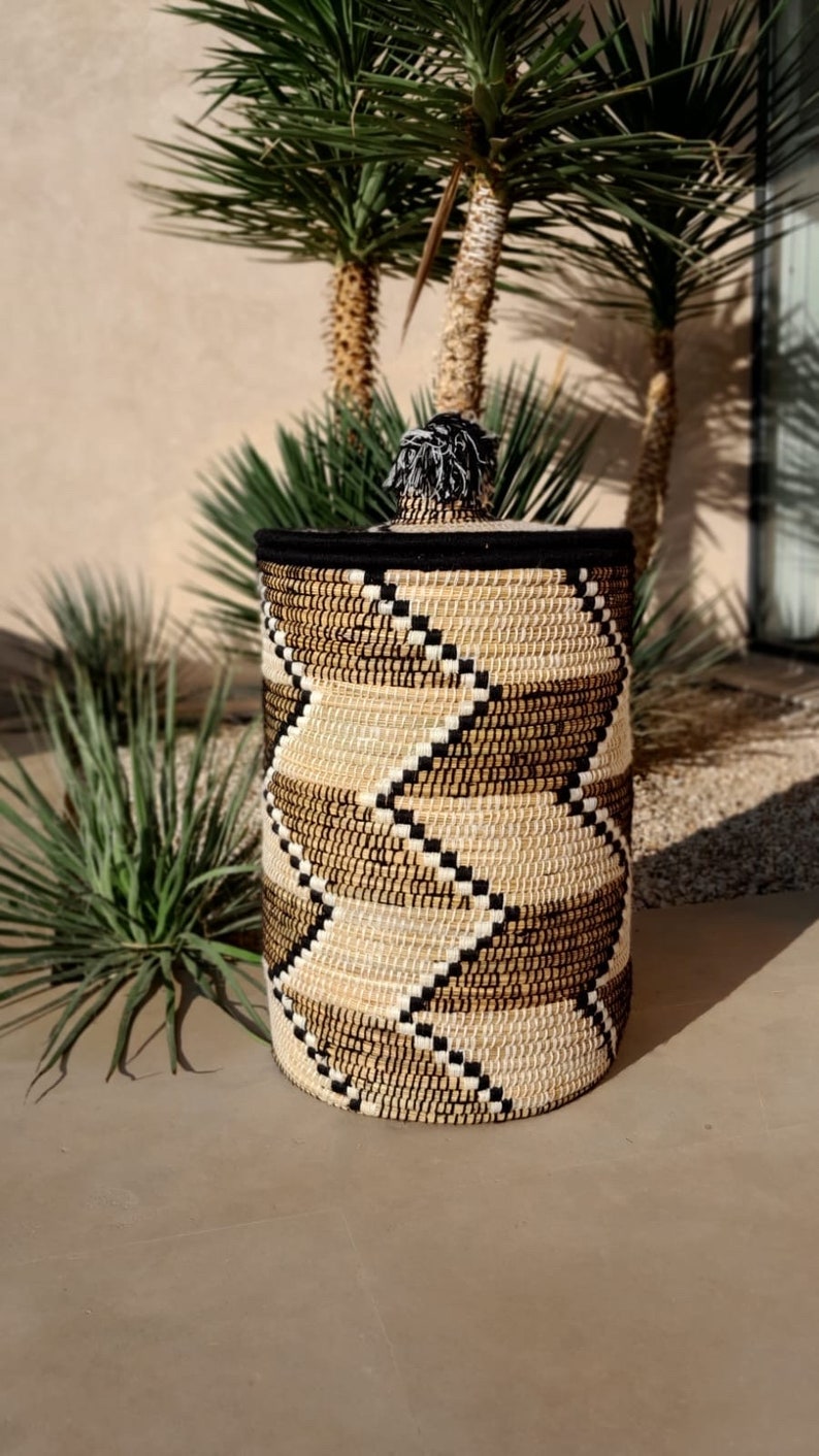 May include: Woven storage basket with a lid. The basket features a geometric pattern in shades of beige, brown, and black. The lid is black with a decorative tassel. The basket is cylindrical and appears to be made of natural fibers.