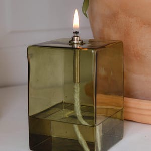 Könnte beinhalten: Eine grüne Glas-Öllampe mit einem Docht und einer Flamme, die im Inneren brennt. Die Lampe steht auf einer weißen Oberfläche.