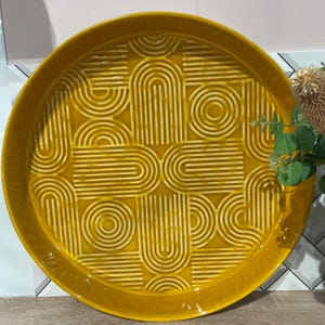 May include: A round, mustard-yellow ceramic serving tray with a raised geometric pattern of arches and lines. The tray has a glossy finish and a slightly raised edge. Decorative item for the home.