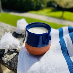 Peut inclure: Un pot de bougie bleu et terracotta avec deux mèches. La bougie est posée sur une serviette rayée blanche et bleue.