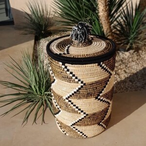 May include: A cylindrical woven basket with a lid. The basket features a geometric pattern in shades of beige, brown, and black. The lid is topped with a black and white tassel. The basket is approximately 12 inches tall and 10 inches in diameter.