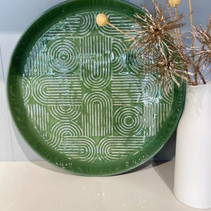 Peut inclure: Un plat de service rond en céramique verte avec un motif géométrique en relief. Le plat présente un motif répétitif de lignes courbes et de cercles. Des tiges florales décoratives dorées sont dans un vase blanc à droite.