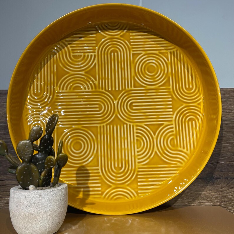 May include: A round, mustard-yellow ceramic tray with a raised geometric pattern of arches and circles. The tray has a glossy finish and a slightly raised edge. A small potted cactus sits to the left of the tray.