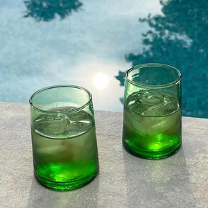 Peut inclure: Deux verres à boire en verre vert remplis de glaçons et d'un liquide clair, posés sur une surface grise. Les verres ont une forme légèrement arrondie et sont en verre épais.