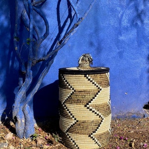 May include: A cylindrical woven basket with a lid. The basket features a repeating chevron pattern in beige, black, and natural tones. The lid is black with a decorative tassel. The background is a vibrant blue wall and a blue tree.