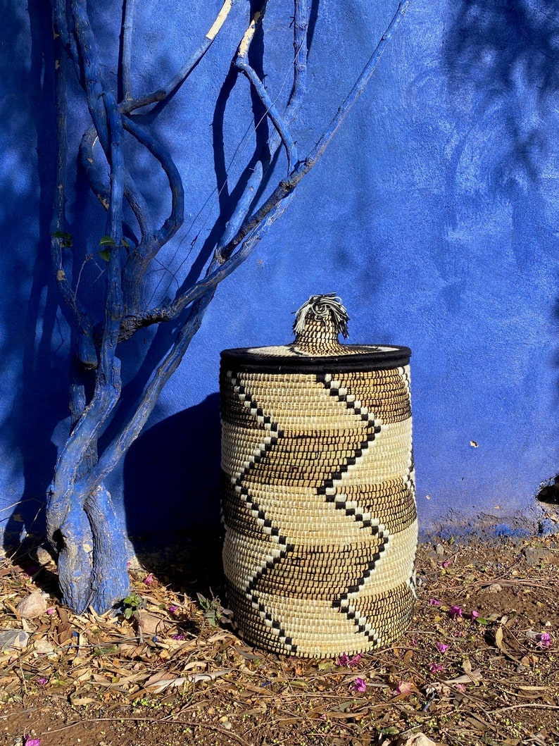 May include: A cylindrical woven basket with a lid. The basket features a repeating chevron pattern in beige, black, and natural tones. The lid is black with a decorative tassel. The background is a vibrant blue wall and a blue tree.