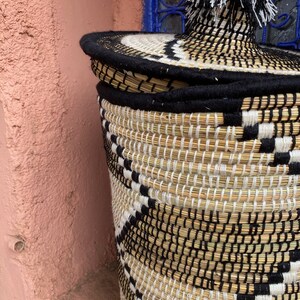 May include: A woven basket with a lid, featuring a black and white geometric pattern. The basket is made of natural fibers and has a black rim and a decorative tassel on top. The background includes a pink wall and a blue window.