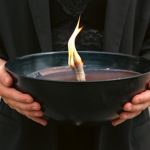 Peut inclure: Une personne tenant un bol noir avec une mèche allumée flottant dans un liquide. La flamme est orange et jaune.