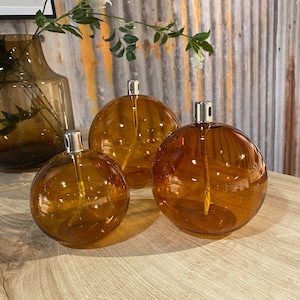 Puede incluir: Tres difusores de aceite esféricos de color ámbar con tapas plateadas se exhiben sobre una superficie de madera. Los difusores varían en tamaño, con un jarrón más grande visible en el fondo. Los difusores de vidrio tienen un diseño clásico y elegante.