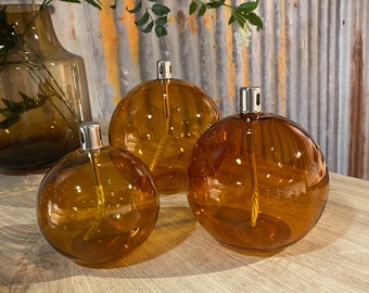 May include: Three amber-coloured, spherical oil diffusers with silver-coloured tops are displayed on a wooden surface. The diffusers vary in size, with a larger vase visible in the background. The glass diffusers have a classic, elegant design.