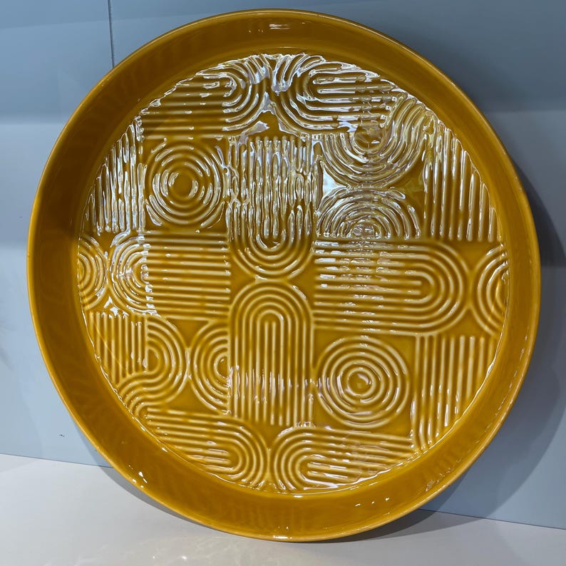 May include: A round, mustard-yellow ceramic serving platter with a raised geometric pattern. The design features repeating arches, circles, and vertical lines. The platter has a slightly raised edge.