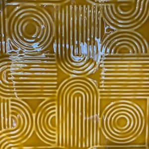 May include: Close-up of a square, mustard-yellow tile with a glossy finish. The tile features a repeating pattern of curved lines, arches, and circles, creating a retro, geometric design. The design is raised, giving the tile a textured appearance.