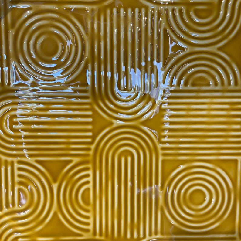 May include: Close-up of a square, mustard-yellow tile with a glossy finish. The tile features a repeating pattern of curved lines, arches, and circles, creating a retro, geometric design. The design is raised, giving the tile a textured appearance.