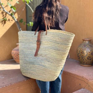 May include: A large, woven straw tote bag with brown leather handles and a buckle closure. The bag is rectangular with a wide opening, suitable for carrying groceries or beach items. The bag is held by a person wearing a black shirt and blue jeans.
