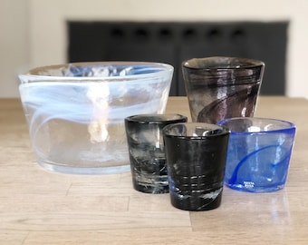 Puede incluir: Una colección de cinco recipientes de vidrio que incluye un cuenco grande transparente con patrones blancos en espiral, un vaso alto de color morado oscuro, dos vasos gris oscuro texturizados y un vaso azul con el texto Kosta Boda.