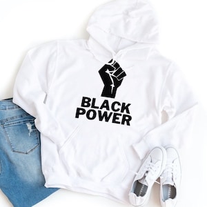 Puede incluir: Sudadera con capucha blanca con un puño negro levantado y el texto "BLACK POWER" impreso en la parte delantera.