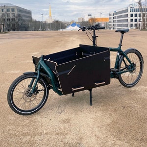Könnte beinhalten: Ein grünes Lastenrad mit einer schwarzen Holzkiste. Das Fahrrad hat einen schwarzen Sattel und einen schwarzen Lenker. Das Fahrrad steht auf einem Schotterplatz.