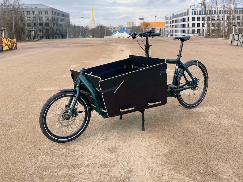 May include: A green cargo bike with a black wooden cargo box. The bike has a black seat and black handlebars. The bike is parked on a gravel surface.
