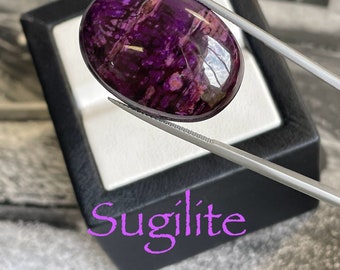 May include: A close-up of a purple and white sugilite gemstone, oval-shaped, with a polished finish. The gemstone is held by tweezers and is in a black velvet box. The word "Sugilite" is written in pink below the box.