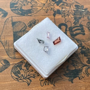 May include: Four faceted gemstones in a white foam-lined box. The gemstones are pink, gray, and a darker shade of pink. The box is on a wooden surface with a floral pattern.