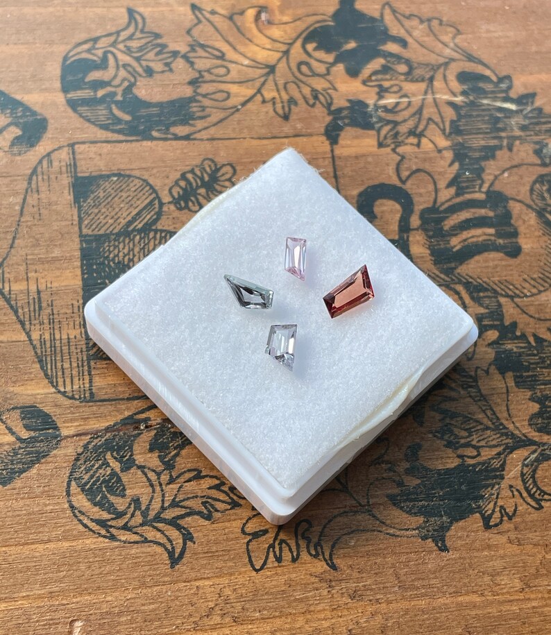 May include: Four faceted gemstones in a white foam-lined box. The gemstones are pink, gray, and a darker shade of pink. The box is on a wooden surface with a floral pattern.