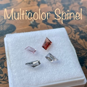 May include: Four loose spinel gemstones in a white box. The gemstones are in various shapes and sizes. The gemstones are pink, red, and clear.