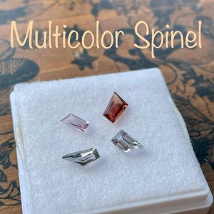 May include: Four loose spinel gemstones in a white box. The gemstones are in various shapes and sizes. The gemstones are pink, red, and clear.