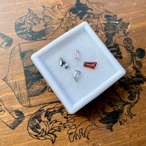 May include: Four small, rectangular gemstones in a white box. The gemstones are a light pink, a dark red, a clear white, and a dark gray.