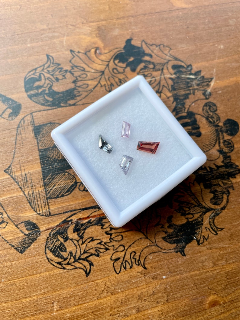 May include: Four small, rectangular gemstones in a white box. The gemstones are a light pink, a dark red, a clear white, and a dark gray.