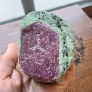 May include: A large, rough, green and pink gemstone with black streaks. The gemstone is a raw, uncut piece of ruby zoisite.