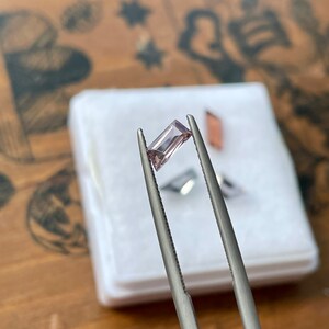 May include: A light purple gemstone held by tweezers. The gemstone is rectangular in shape and has a slight shimmer.