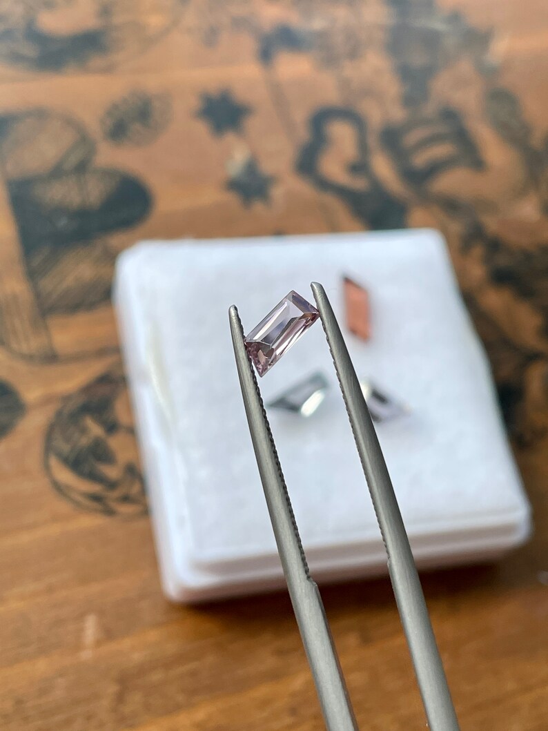 May include: A light purple gemstone held by tweezers. The gemstone is rectangular in shape and has a slight shimmer.