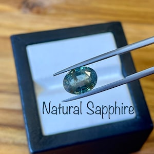 Può includere: Uno zaffiro naturale verde tenuto da una pinza in una scatola per anelli nera con interno bianco. La scatola ha la scritta "Natural Sapphire" stampata sul davanti.