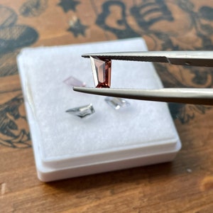 May include: A small, rectangular, dark red gemstone held by tweezers over a white box. The gemstone is a cut and polished piece of raw material.
