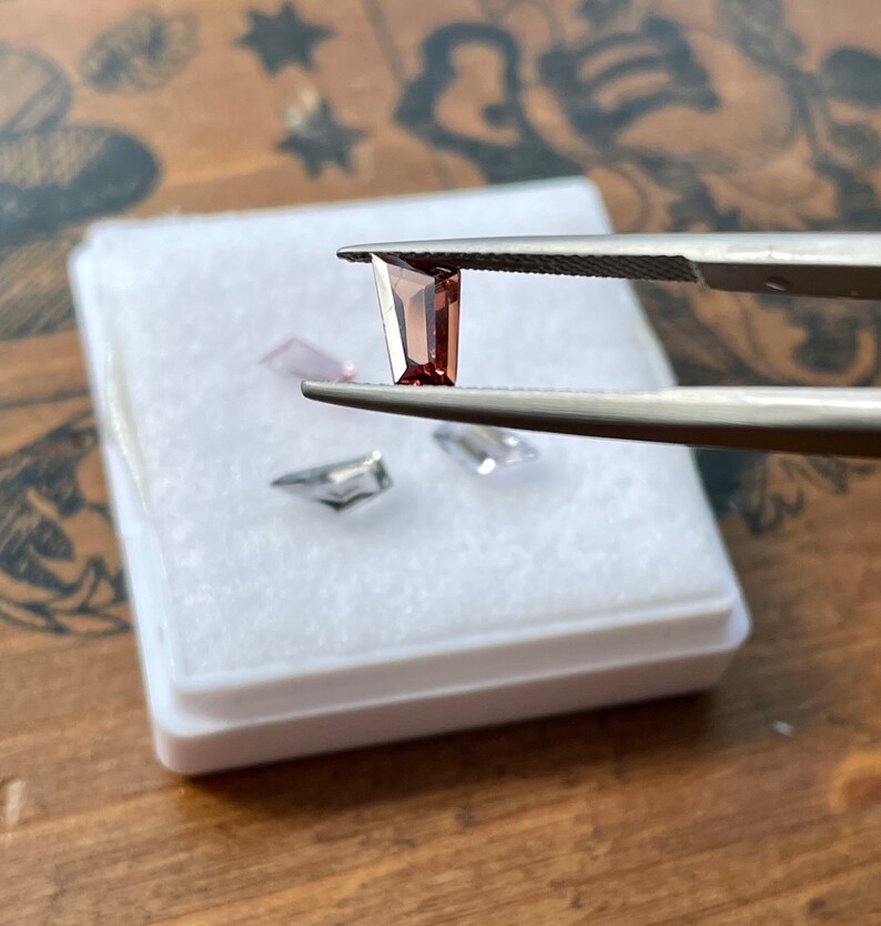 May include: A small, rectangular, dark red gemstone held by tweezers over a white box. The gemstone is a cut and polished piece of raw material.