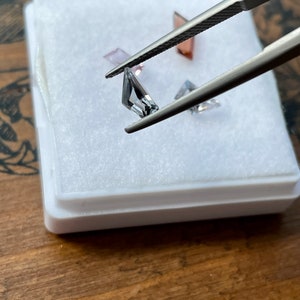May include: A set of three faceted gemstones in a white box. The gemstones are a light pink, a clear gray, and a darker gray. The gemstones are held by tweezers.