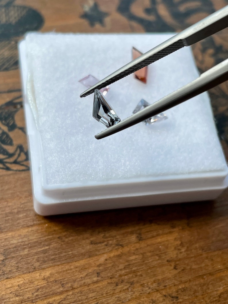 May include: A set of three faceted gemstones in a white box. The gemstones are a light pink, a clear gray, and a darker gray. The gemstones are held by tweezers.