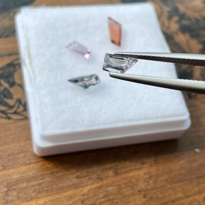 May include: Four small, rectangular, faceted gemstones in a white box. The gemstones are pink, clear, and a light purple. The gemstones are being held by tweezers.