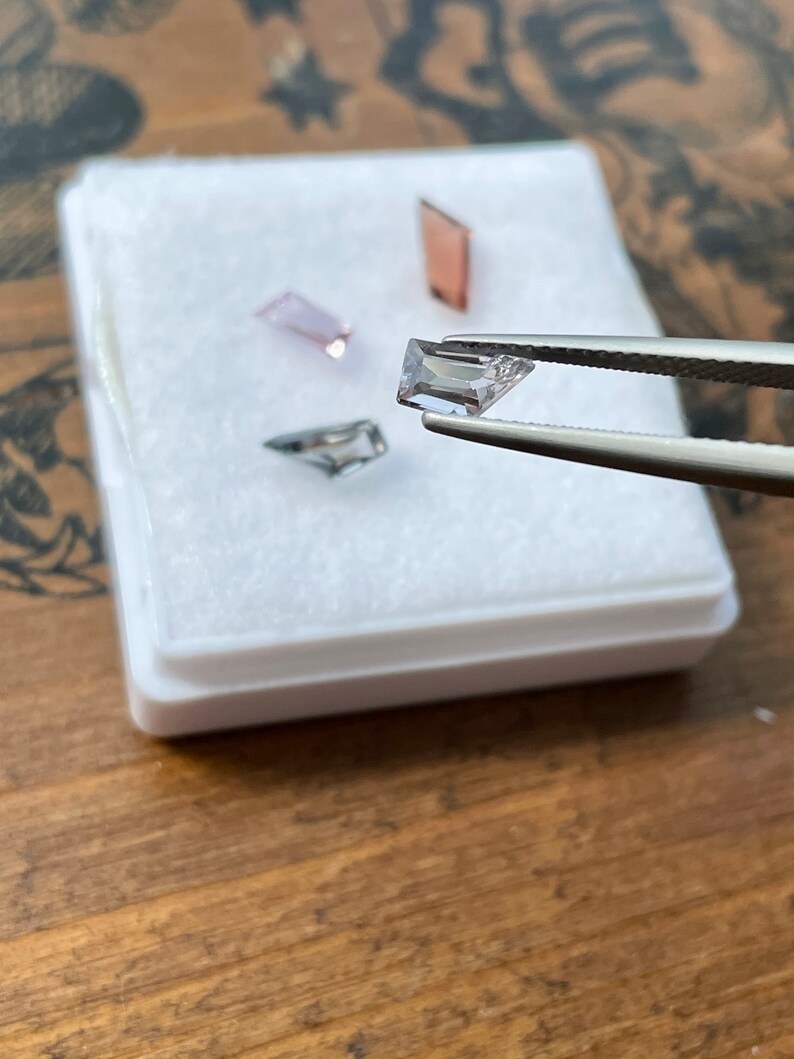 May include: Four small, rectangular, faceted gemstones in a white box. The gemstones are pink, clear, and a light purple. The gemstones are being held by tweezers.