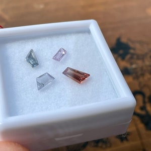May include: Four loose gemstones in a white box. The gemstones are a light blue, a light pink, a light purple, and a light brown.