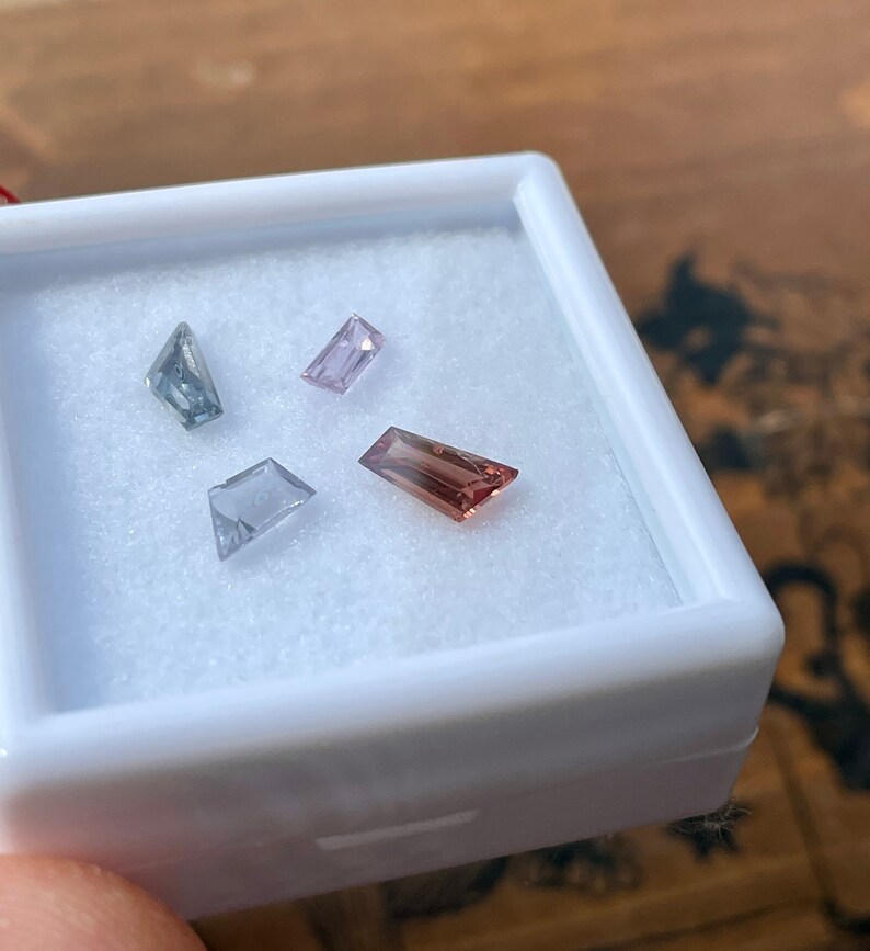 May include: Four loose gemstones in a white box. The gemstones are a light blue, a light pink, a light purple, and a light brown.