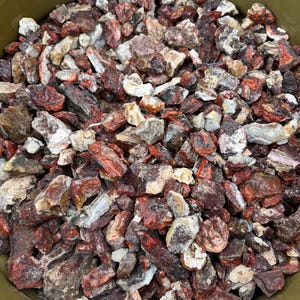 May include: A close-up view of a pile of raw, rough-cut rocks. The rocks are a mix of colors, including deep reds, browns, and off-white. The texture of the rocks appears to be coarse and uneven, suggesting they are natural stones. The rocks are likely used for landscaping or decorative purposes.