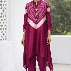 Può includere: Un completo tunica e pantaloni color magenta. La tunica presenta un motivo a chevron bianco e magenta, con maniche lunghe e orlo asimmetrico. I pantaloni sono dello stesso colore magenta. Viene indossata una collana d'argento.