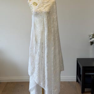 Pu&ograve; includere: Una lunga stola in tessuto bianco semi-trasparente caratterizzata da un intricato ricamo floreale tono su tono su tutta la superficie. Il materiale leggero cade con eleganza, mostrando delicati motivi botanici e un bordo smerlato.